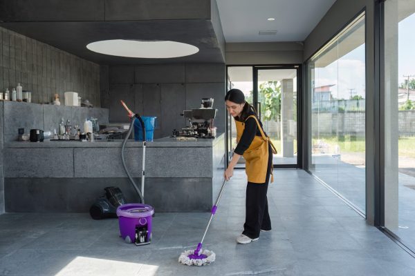 A female employee in a modern coffee shop cleans the floor with a mop and bucket, ensuring a tidy and welcoming environment.