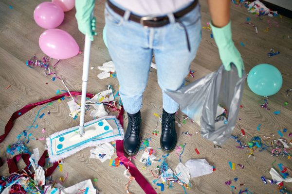Woman with pushbroom cleaning mess of floor in room after party confetti, morning after party celebration, housework, cleaning service